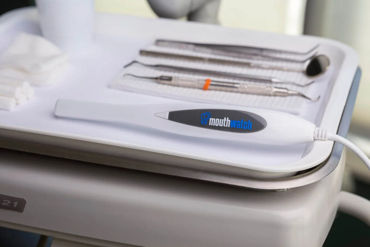 Close-up of a MouthWatch intraoral camera resting on a white dental tray alongside various dental instruments, including a mirror and scaler, in a clinical setting.
