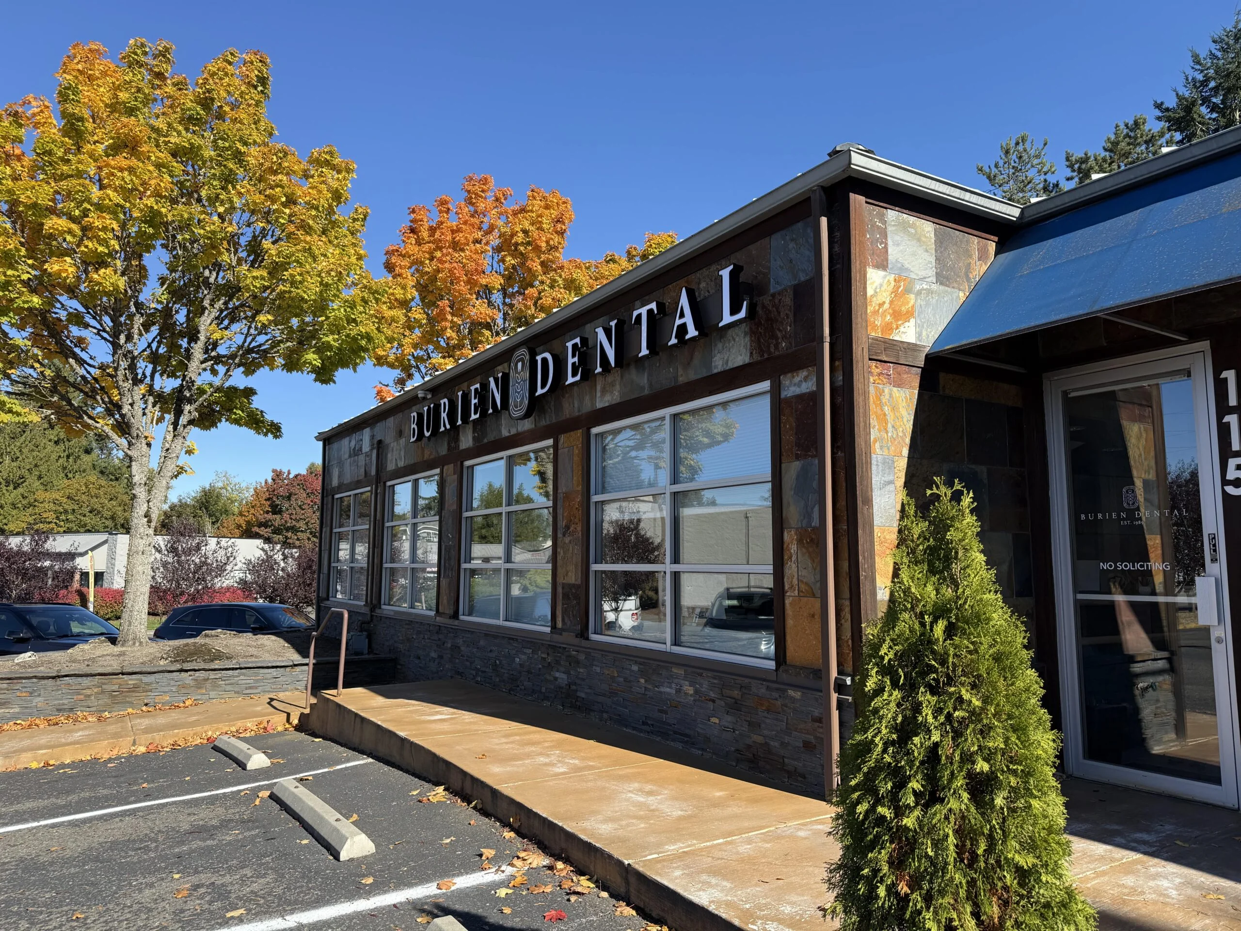 Entrance to the Burien Dental building on a sunny day, with autumn trees in the background.